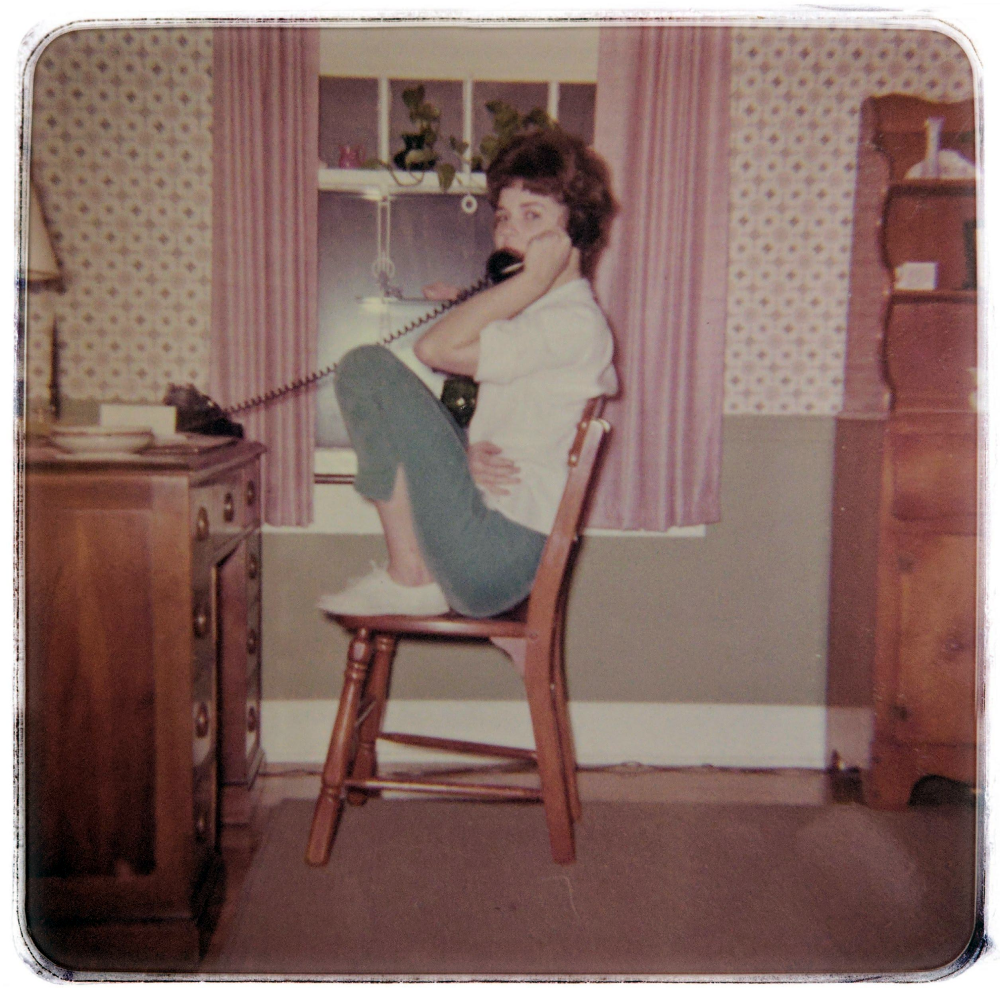 A person sits on a wooden chair with their knees pulled up to their chest while talking on a telephone.