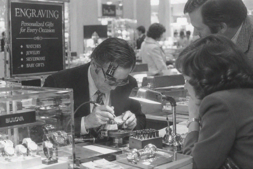 A man wearing a jeweler’s loupe engraves a watch for a customer at a store counter.