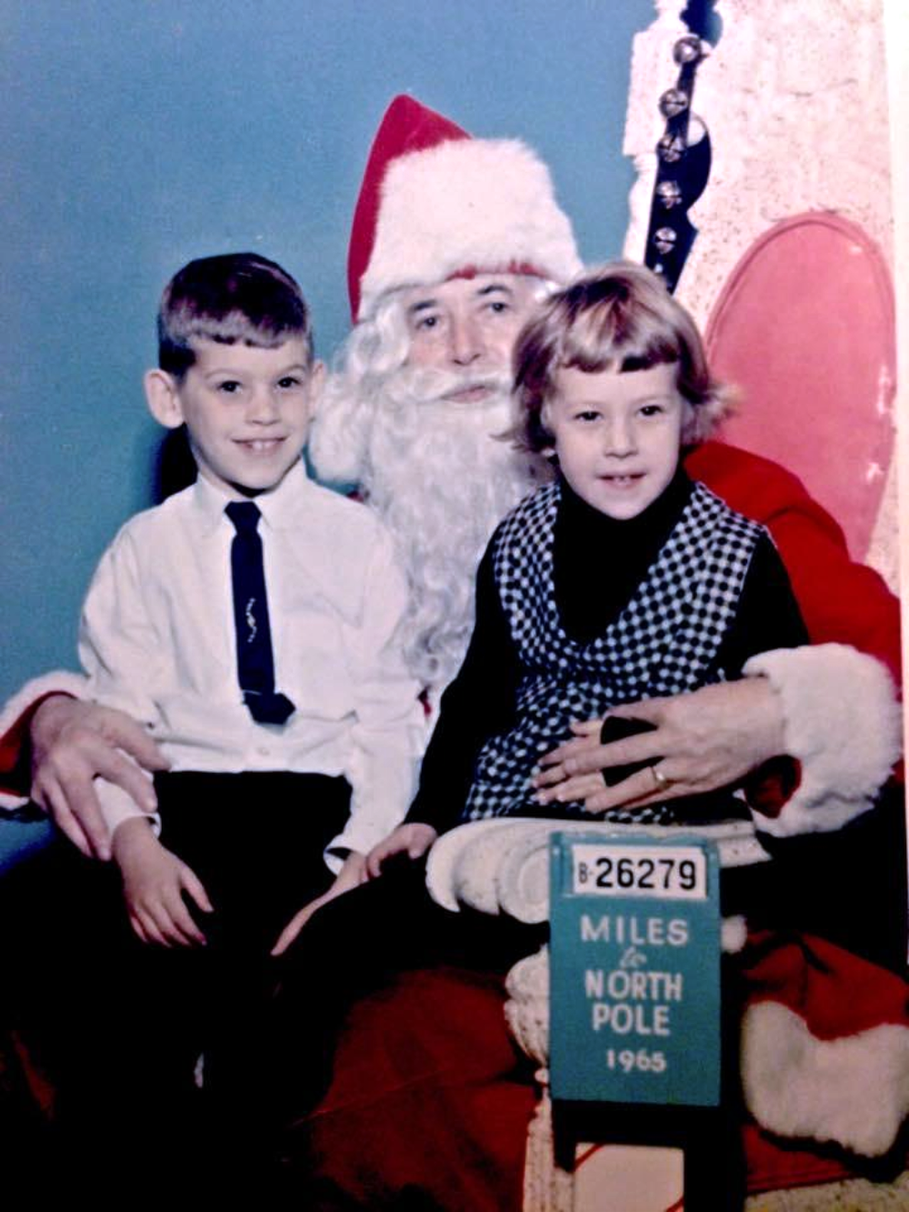 A young boy and girl sitting on Santa Claus’s lap, next to a sign reading “26279 Miles to North Pole 1965.”