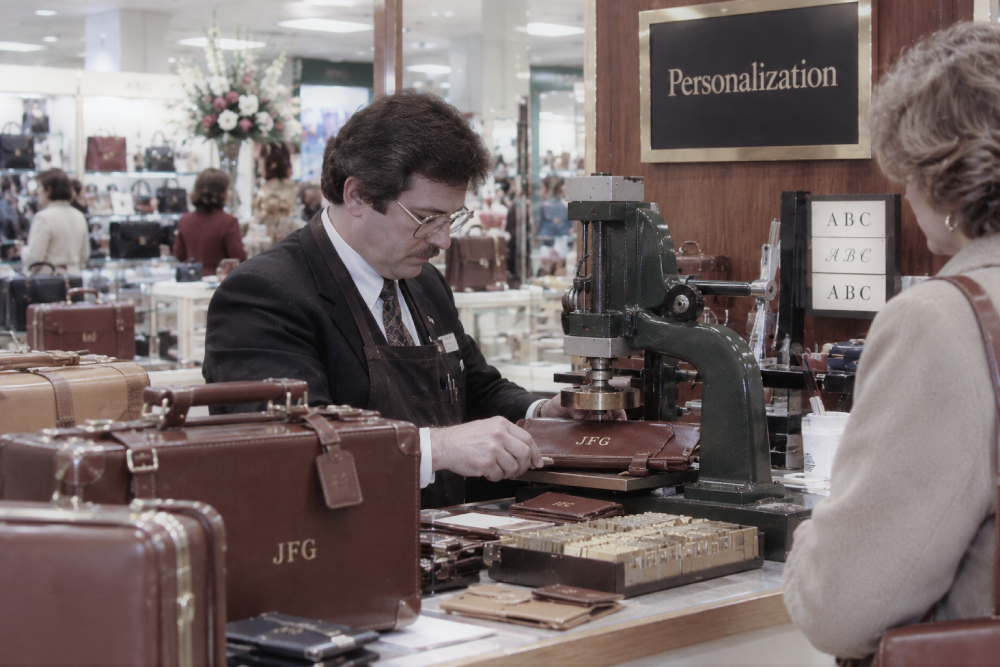A shop employee uses a machine to emboss the initials “JFG” onto a leather item for a customer at a store personalization counter.