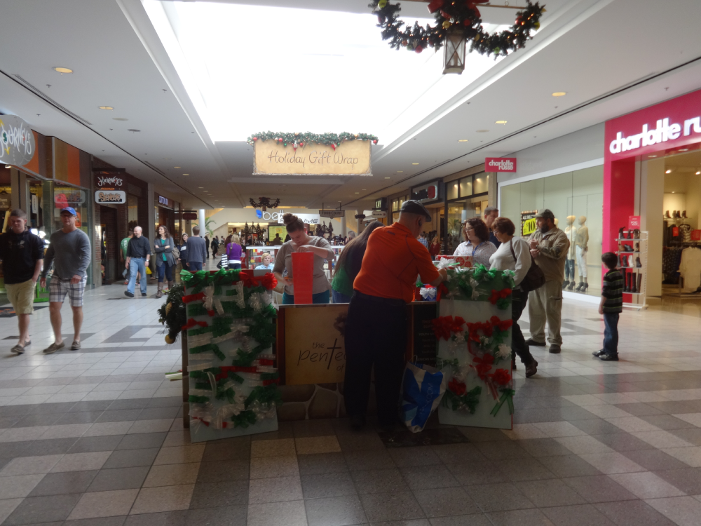 A holiday gift wrapping station in a shopping mall with people standing around a kiosk decorated with festive garland.