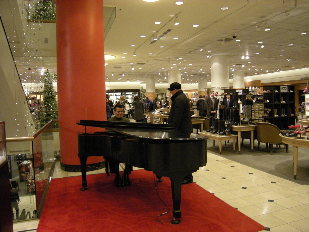 A person playing a black grand piano on a red rug in a department store.