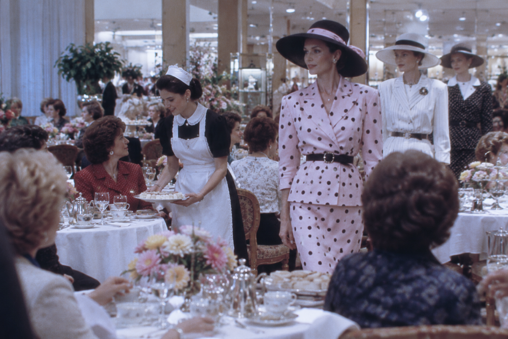 A woman in a pink polka-dot suit and wide-brimmed hat walks through a crowded dining room as a server in a white apron offers a tray of food.