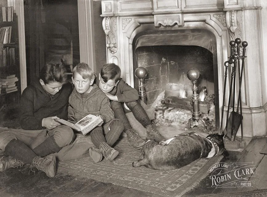 Three boys sit on a rug in front of a fireplace reading a book together, with a dog lying at their feet.