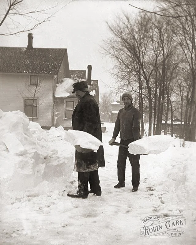 Two men in winter coats shovel deep snow in front of a house.