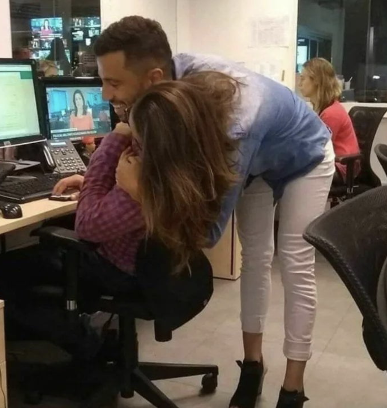 A man leaning over a desk to embrace a woman sitting in an office chair.