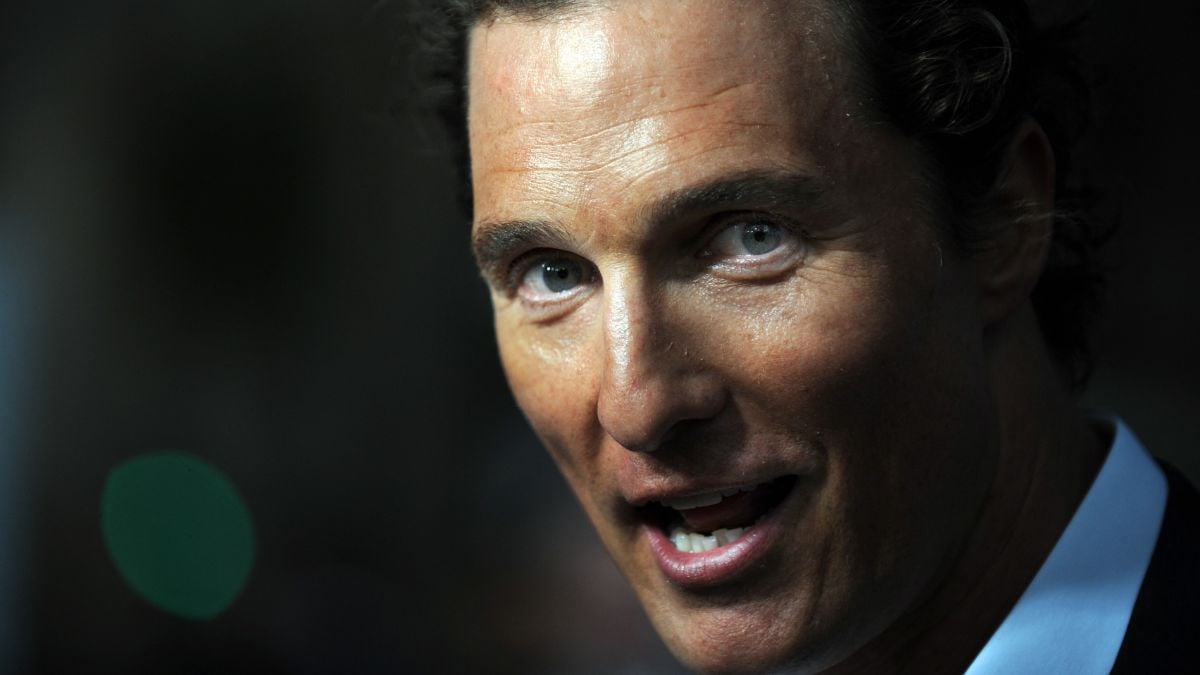 A man with light eyes and curly brown hair is speaking, looking slightly past the camera. The background is blurry and dark, with a green light visible in the distance.