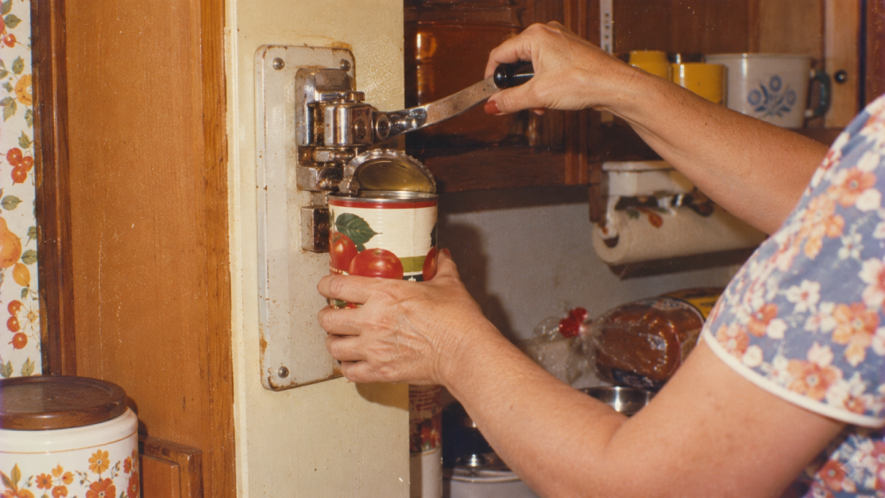 A person using a wall-mounted hand crank can opener to open a tin can decorated with images of tomatoes.