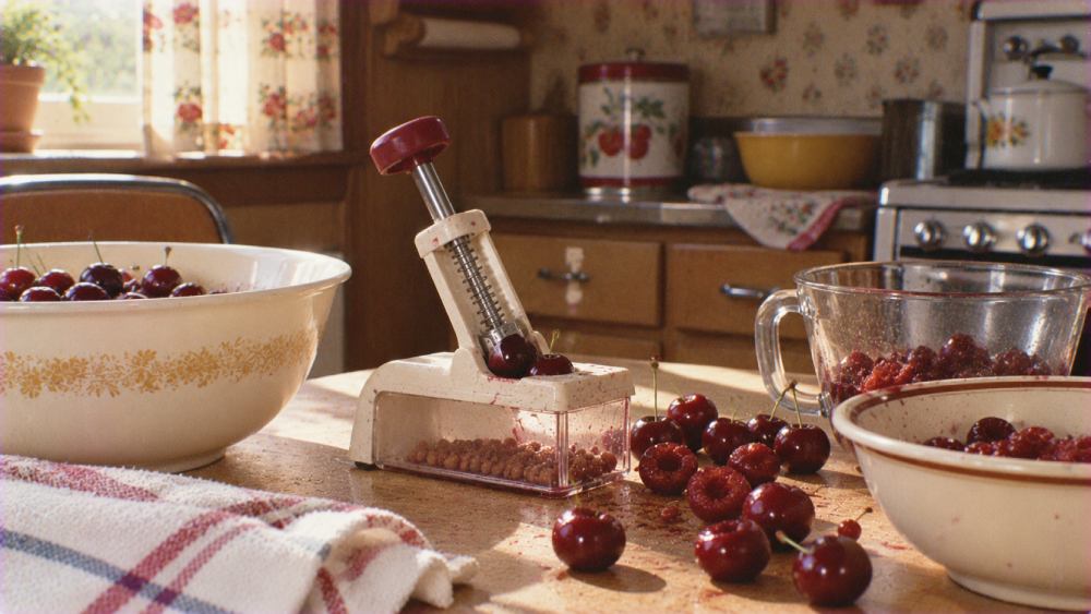 A cherry pitter on a kitchen table surrounded by bowls of fresh cherries and pits.