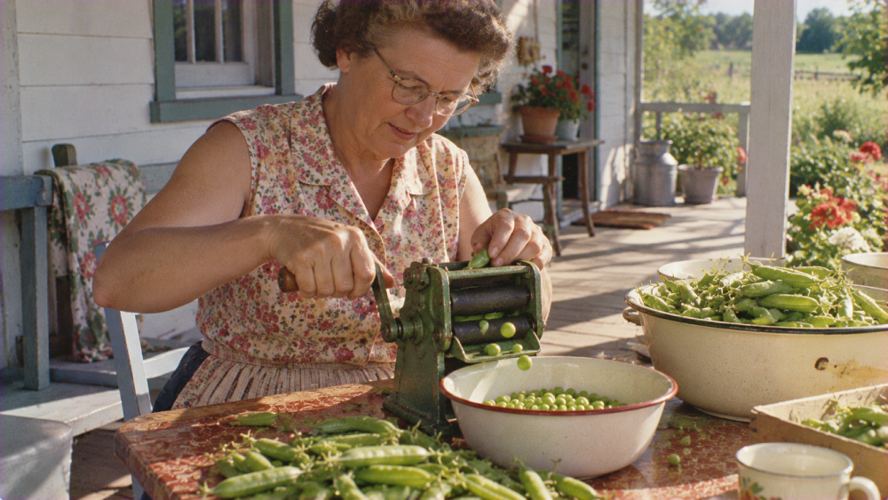 A woman on a porch uses a hand-cranked machine to shell peas into a bowl.