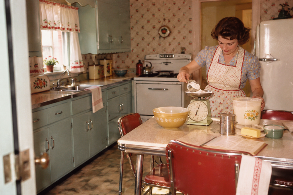 A woman in a floral dress and apron measuring flour with a scale at a kitchen table.