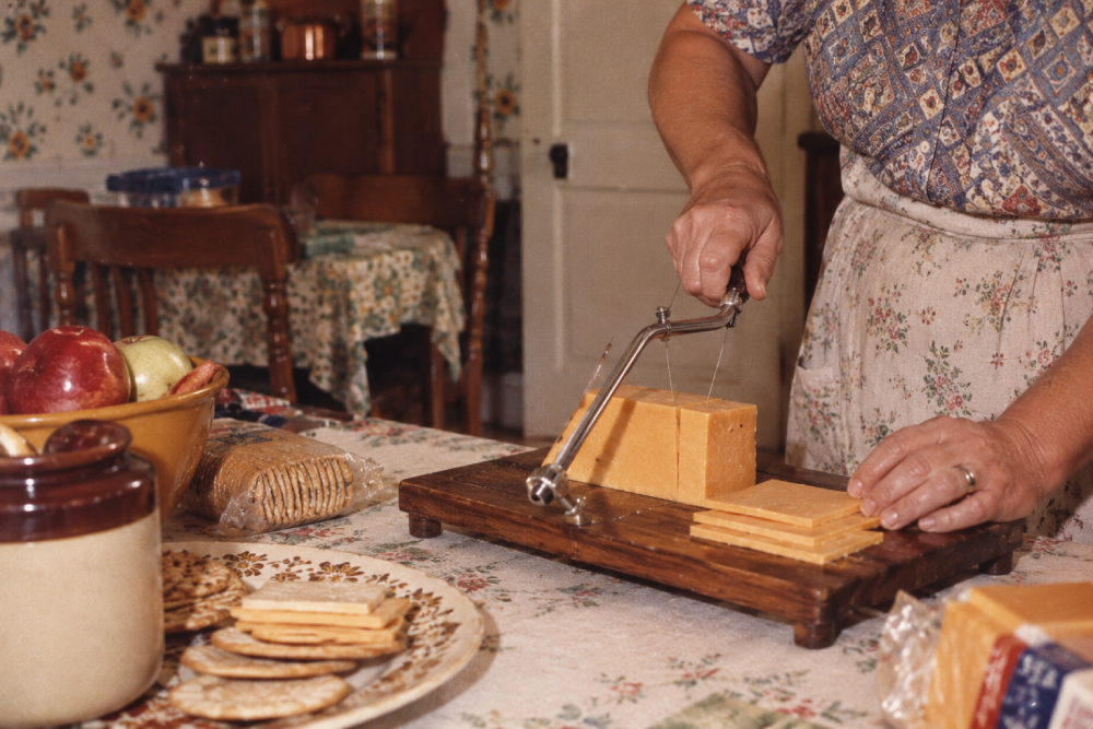 A person uses a wire cheese slicer to cut a block of cheddar cheese on a wooden board.