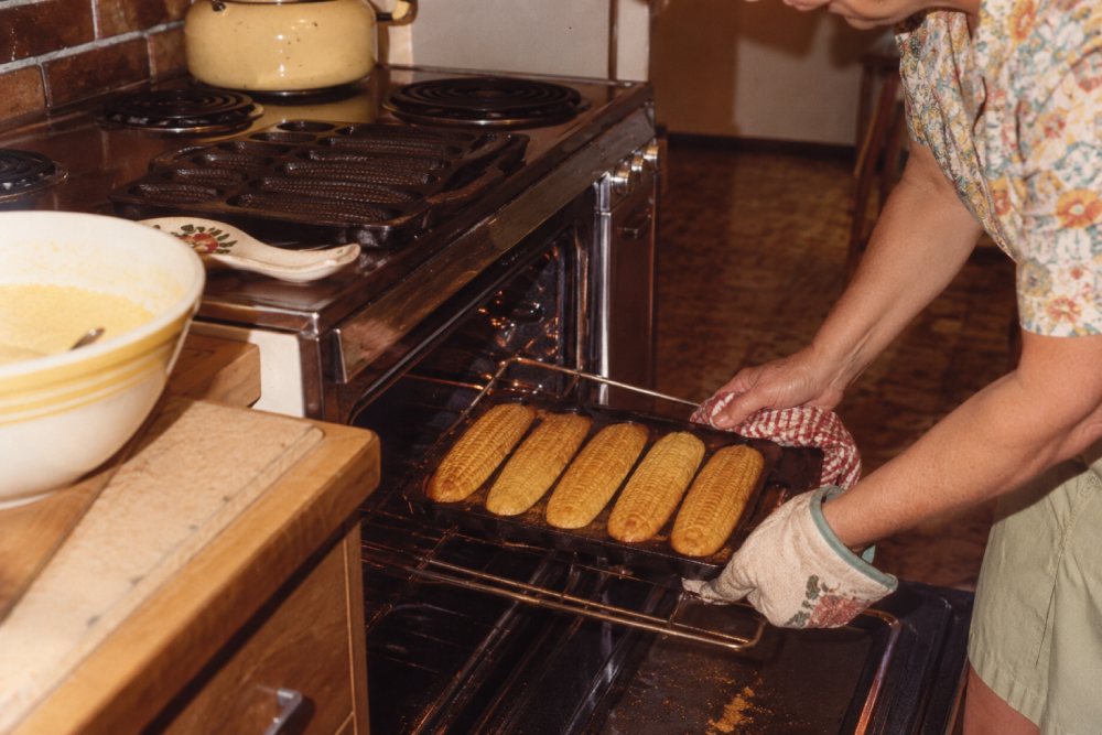 A person wearing an oven mitt slides a pan of freshly baked corn-shaped cornbread into an oven.