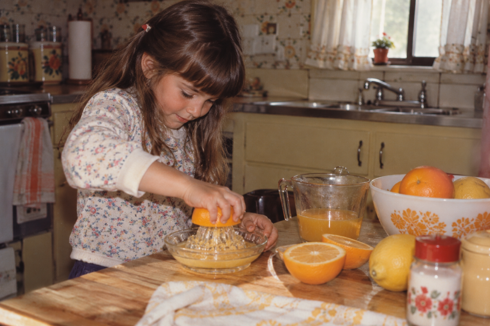 A young girl in a floral shirt juices an orange by hand into a glass bowl on a kitchen table.