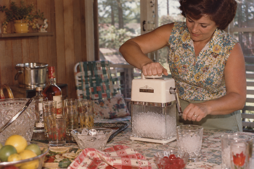 A woman in a floral sleeveless top uses a vintage Ice-O-Mat hand-crank ice crusher at a table set with glassware and citrus.