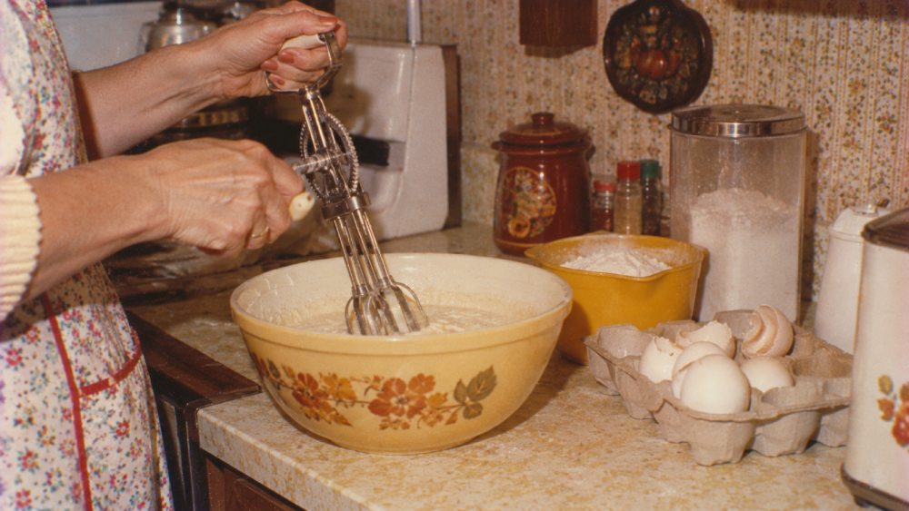 A person uses a handheld rotary egg beater to mix batter in a floral bowl on a kitchen counter.