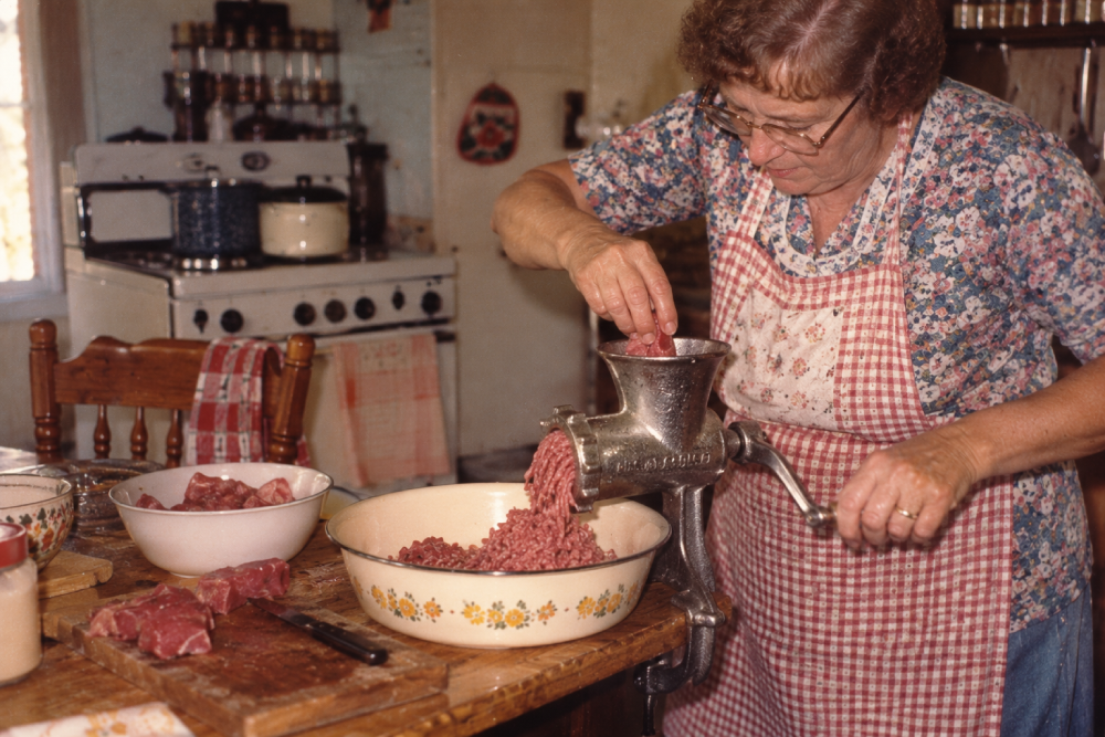 A woman in a floral shirt and plaid apron uses a vintage hand-crank meat grinder to process meat into a bowl.