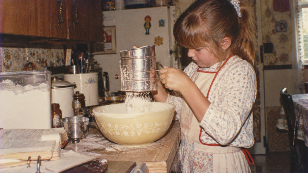 A young girl wearing an apron uses a hand-crank sifter to dust flour into a large mixing bowl on a kitchen counter.