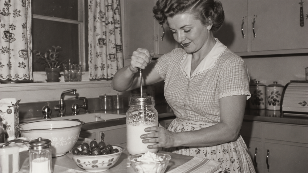 A woman in a kitchen uses a hand-cranked beater to whip cream in a glass jar.