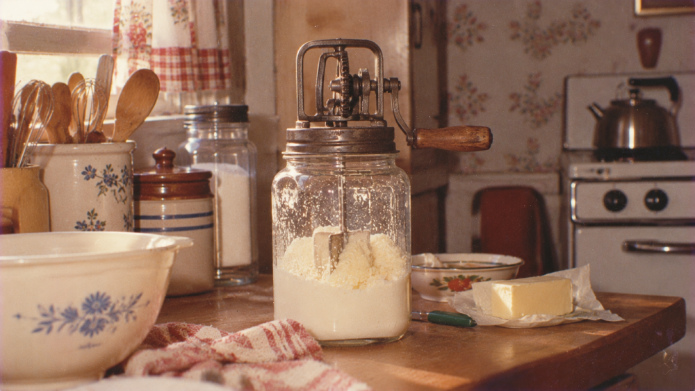 A vintage hand-cranked butter churn sits on a kitchen counter next to a bowl of flour and a stick of butter.