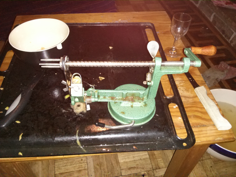 A green metal apple peeler sitting on a black tray on a wooden table.