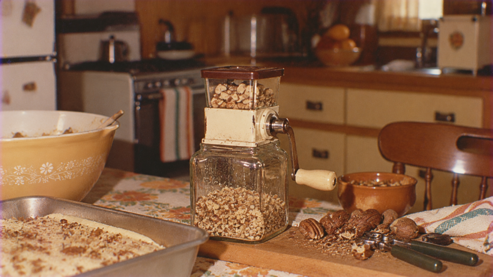 A hand-cranked nut grinder sits on a wooden cutting board next to a metal baking pan, a small bowl of nuts, and a nutcracker on a kitchen table.