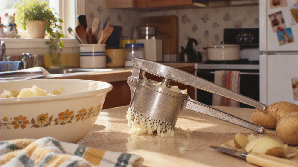 A metal potato ricer filled with riced potatoes sits on a kitchen counter next to a bowl of mashed potatoes and whole potatoes.