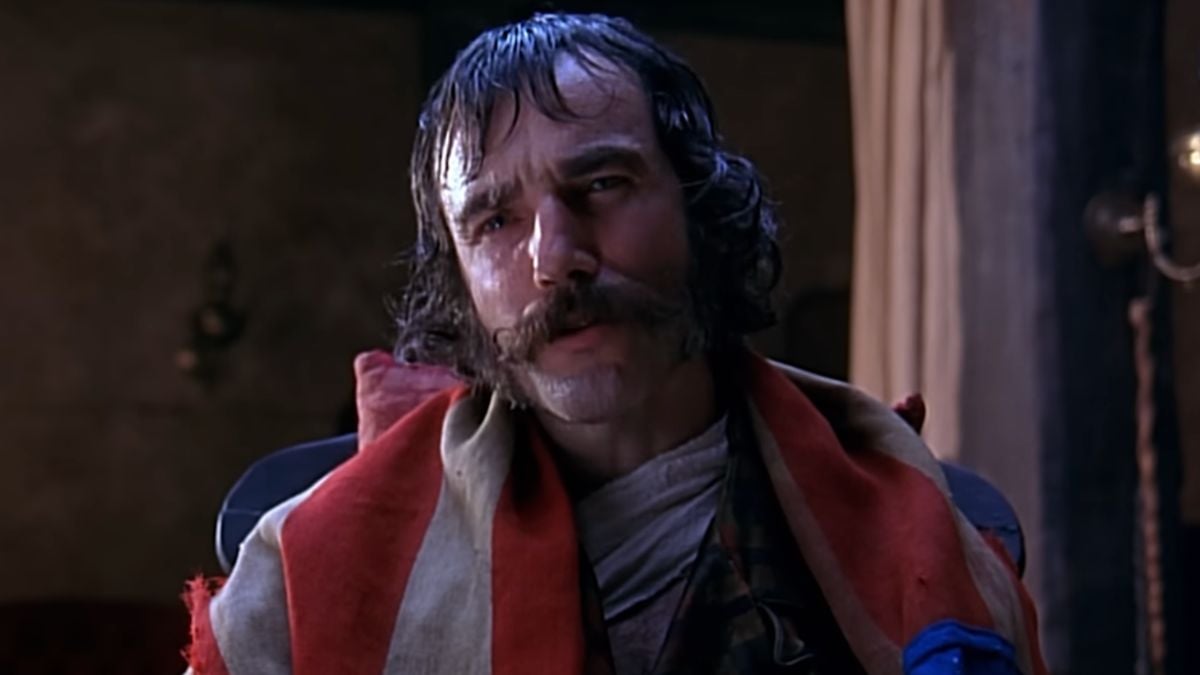 A man with thick mustache and mutton-chop sideburns sits indoors, draped in a flag, looking serious and intense. The background is dimly lit with a curtain and part of a wall visible.