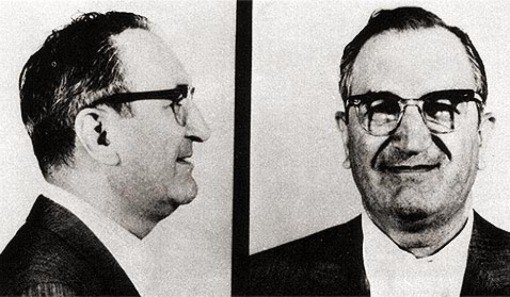 Black-and-white mugshot of a man with glasses, showing his profile and front view.