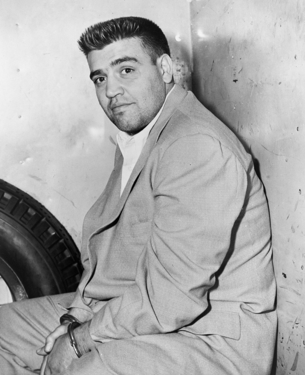 A man in a suit and handcuffs sits against a wall next to a large tire.