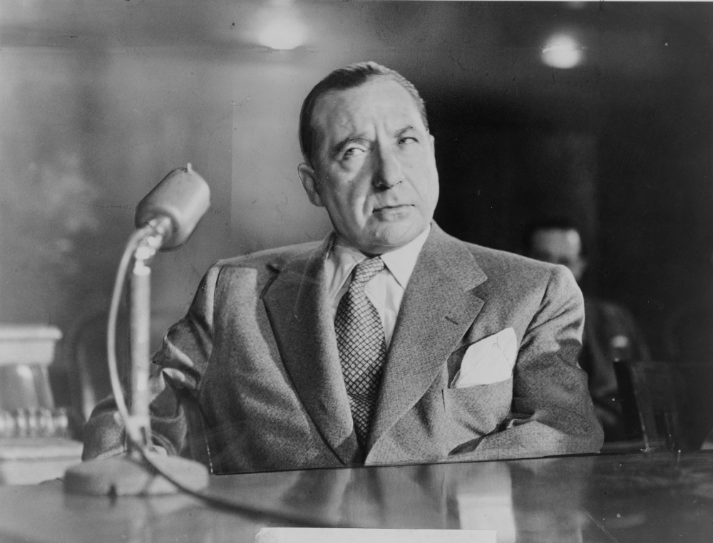 A man in a suit and tie sits at a table speaking into a microphone.