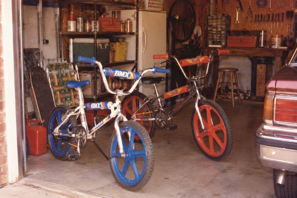 Two BMX bikes with colorful red and blue wheels stand in a garage near a car. The garage has tools, a fridge, a skateboard, and folding chairs visible in the background.