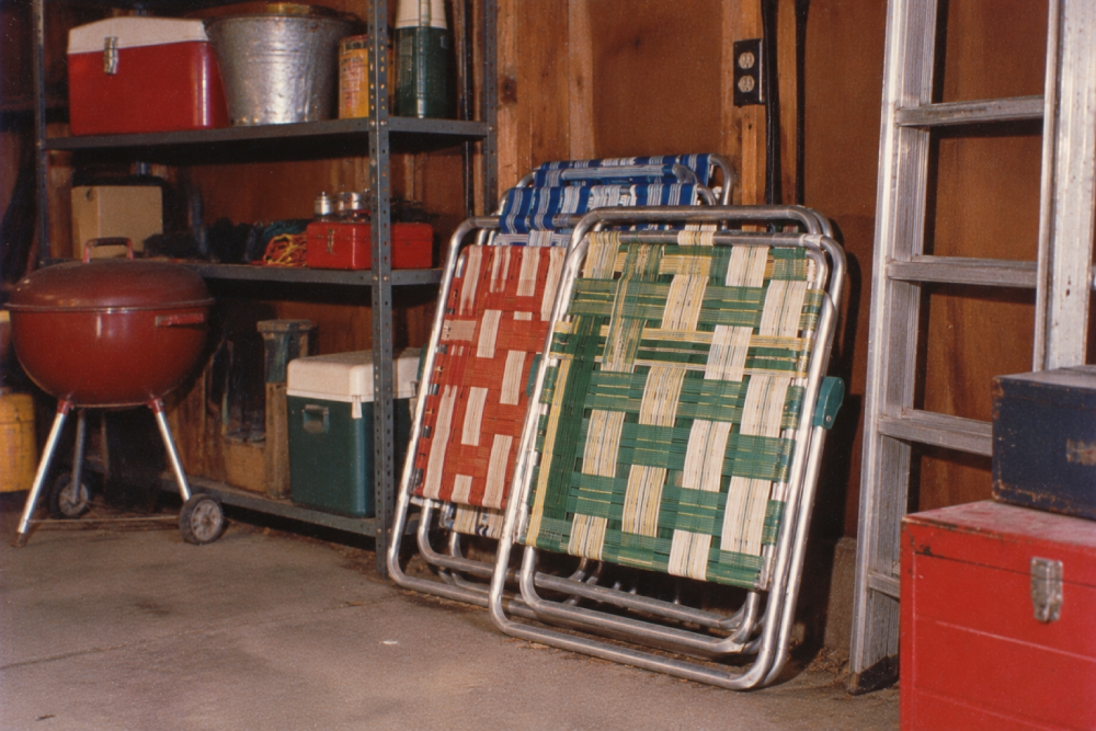 Two folding lawn chairs, one with green and one with red woven webbing, lean against a wooden wall in a garage. Nearby are a metal shelf with coolers, a red grill, a ladder, and a red toolbox.