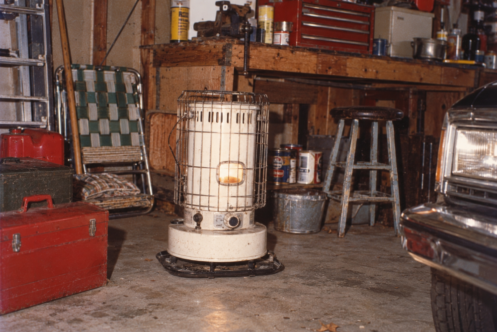 A lit kerosene heater sits on a garage floor surrounded by a toolbox, lawn chair, stool, and various tools, with a car bumper visible on the right.