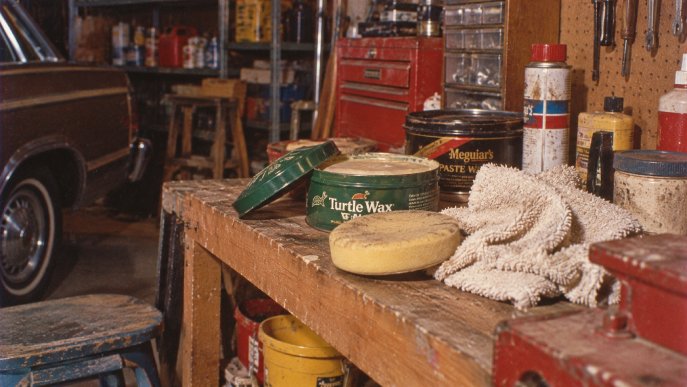 A cluttered garage workbench with Turtle Wax, a yellow sponge, rags, and various car care products. A classic car is partially visible to the left, with tools and supplies scattered around.