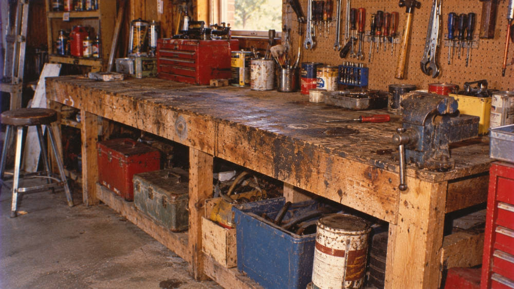 A wooden workbench in a workshop, cluttered with tools, paint cans, toolboxes, a vise, and various supplies. Pegboard above holds hanging tools. A stool and more toolboxes sit beneath the workbench.