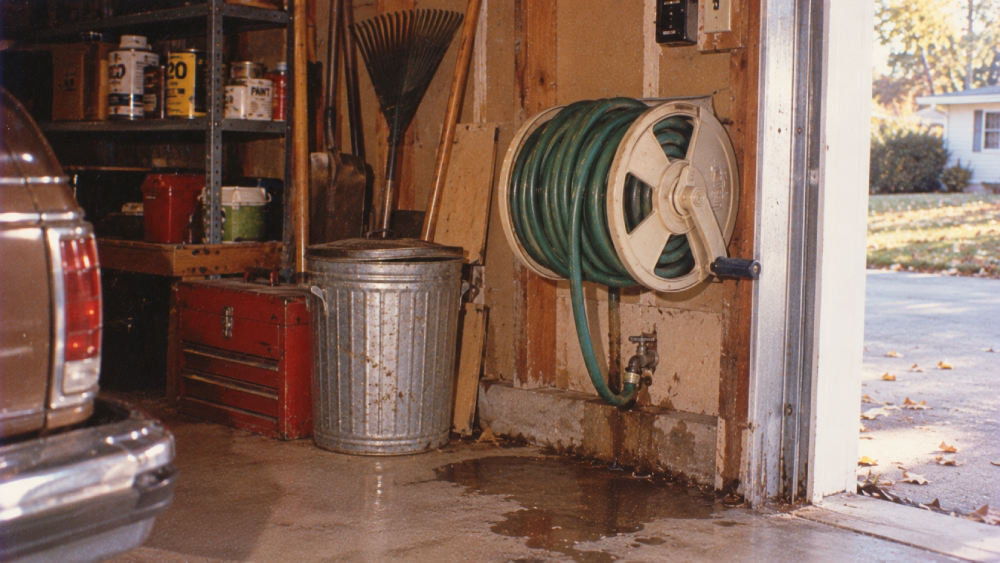 A metal trash can and garden hose reel hang on a garage wall. Tools and paint cans are on shelves. Part of a brown car is visible, and sunlight shines in from an open garage door onto a wet concrete floor.