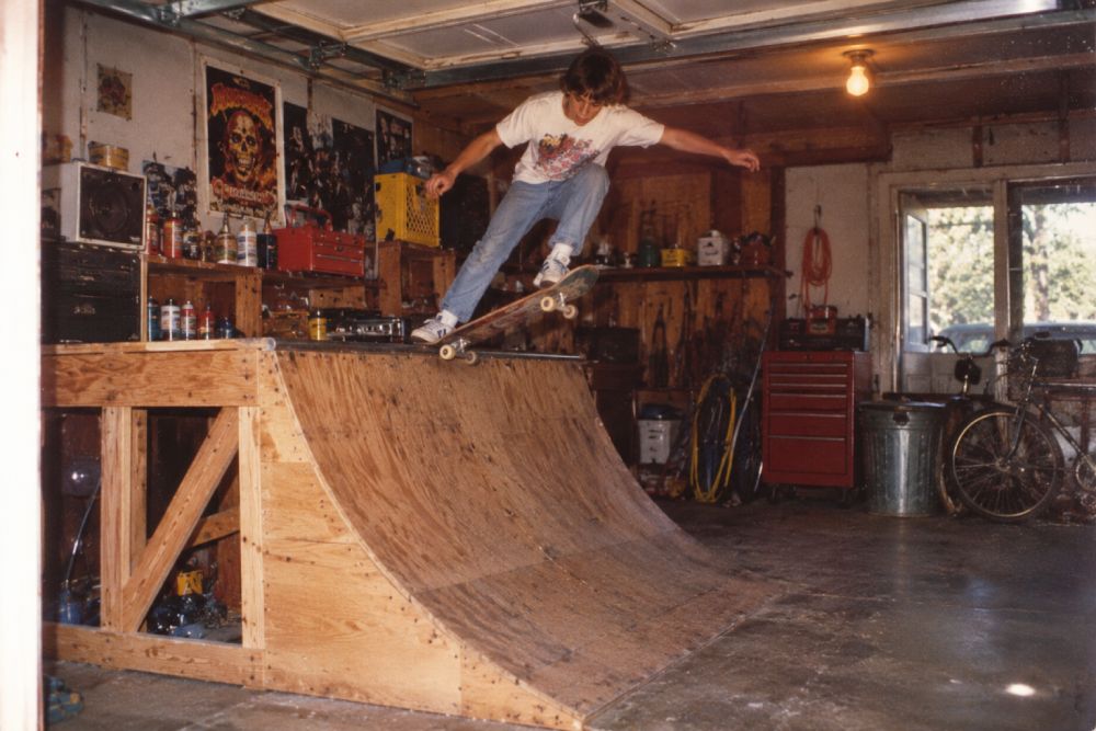 A person skateboards on a homemade wooden ramp inside a garage filled with posters, tools, and a bicycle near a window, with sunlight shining in.