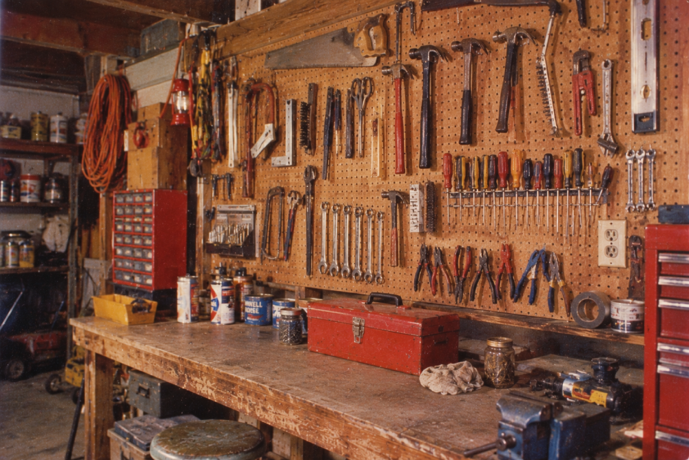 A wooden workbench with a red toolbox sits below a pegboard full of organized tools like hammers, wrenches, and screwdrivers in a workshop. Painted cans and other supplies are also on the bench.