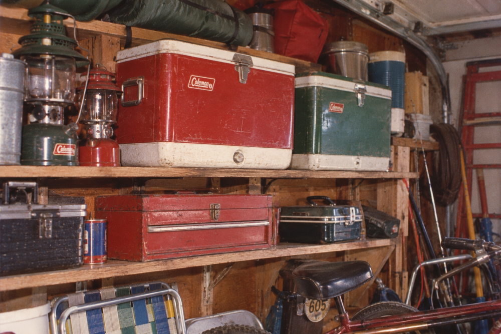 Shelves in a garage hold vintage coolers, lanterns, toolboxes, folding chairs, and other camping gear. A bicycle is partially visible in the foreground. The area looks organized but packed with supplies.