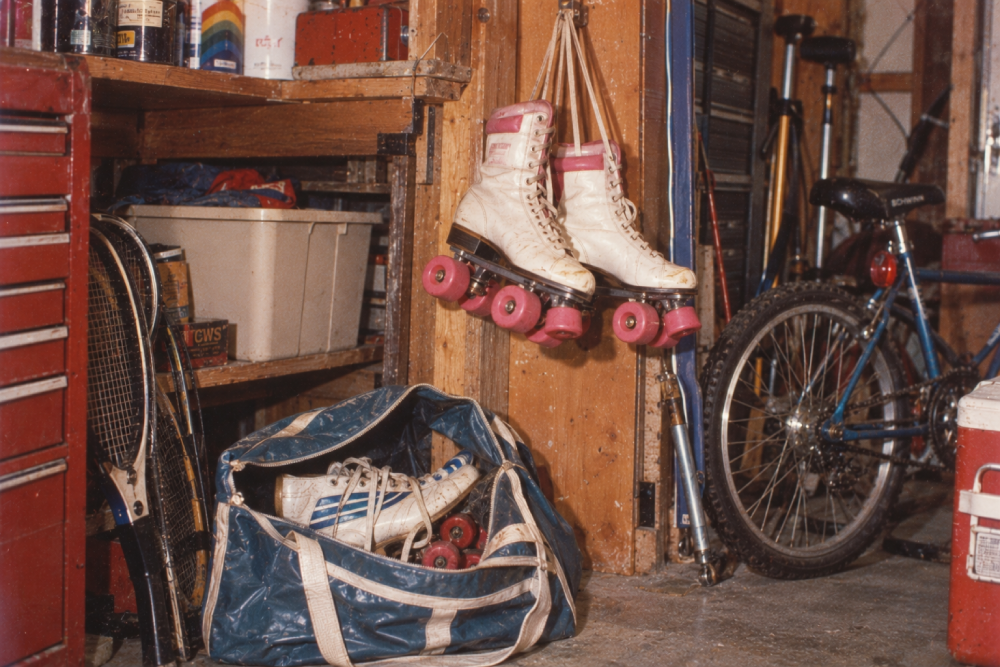 A pair of white roller skates hang on a wall near a blue bike, a sports bag with another pair of skates, tennis rackets, tools, and shelves in a cluttered garage.