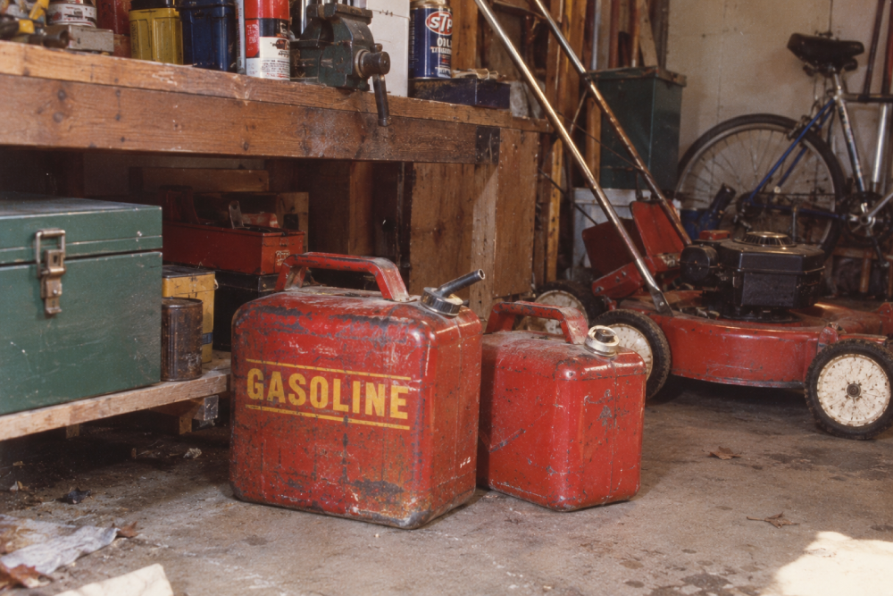 Two red gasoline cans sit on a garage floor near a red lawnmower, green toolbox, bicycle, and shelves with various tools and containers.