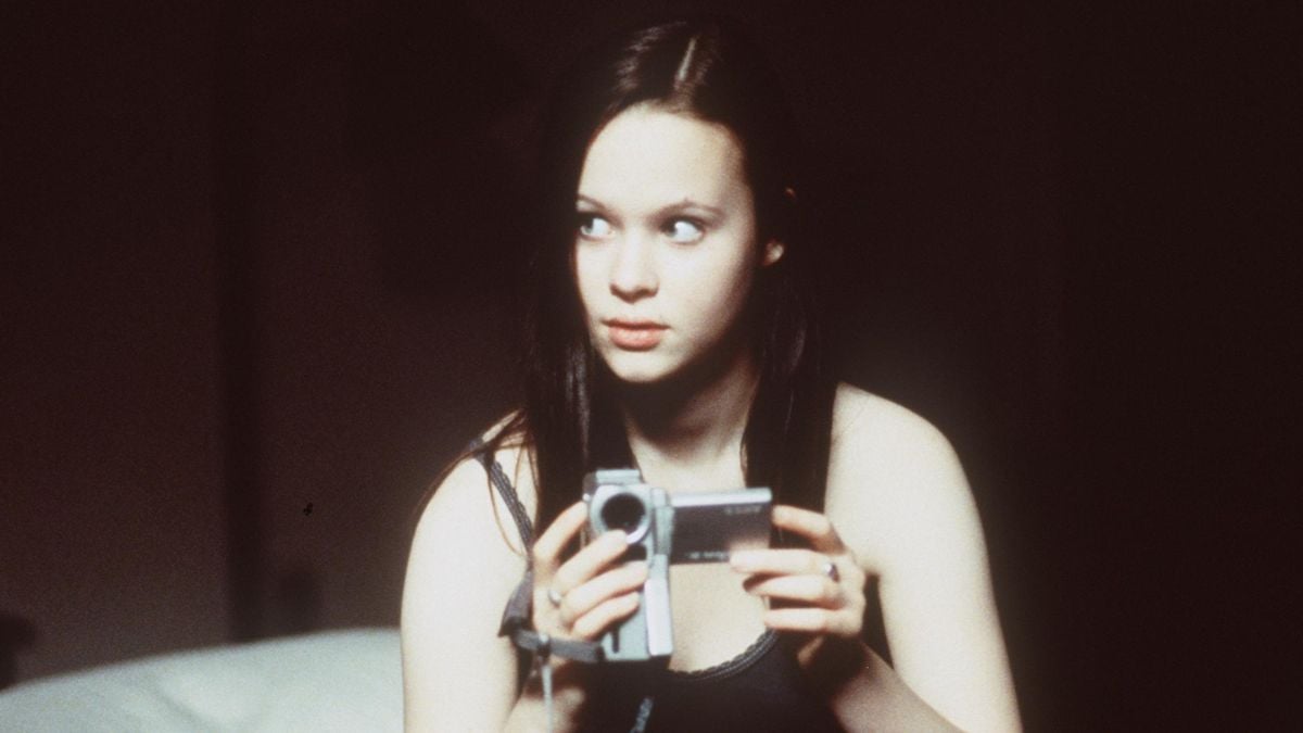 A young woman with long dark hair sits indoors, holding a small handheld video camera, looking off to the side with a serious expression.