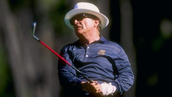 A golfer wearing a white hat, sunglasses, and a blue shirt swings a golf club outdoors, with a blurred green background.