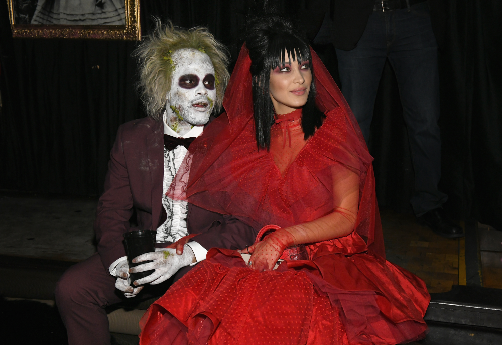Two people in Beetlejuice and Lydia Deetz costumes sit together, the man in a messy wig and white face paint and the woman in a red bridal gown and veil.
