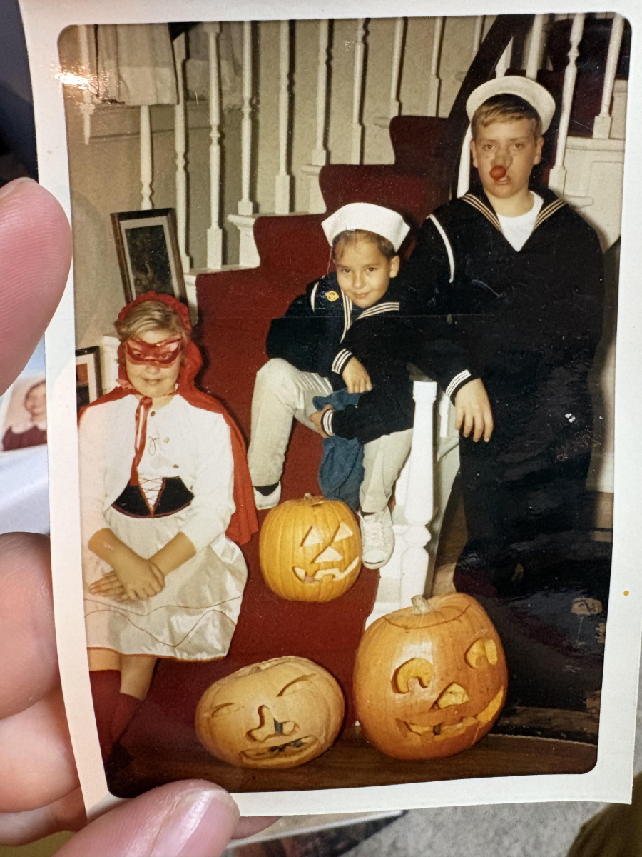Three children in Halloween costumes pose on stairs with three carved pumpkins. Two kids wear sailor outfits; one girl wears a red cape and mask. The photo shows part of a staircase and red carpet.