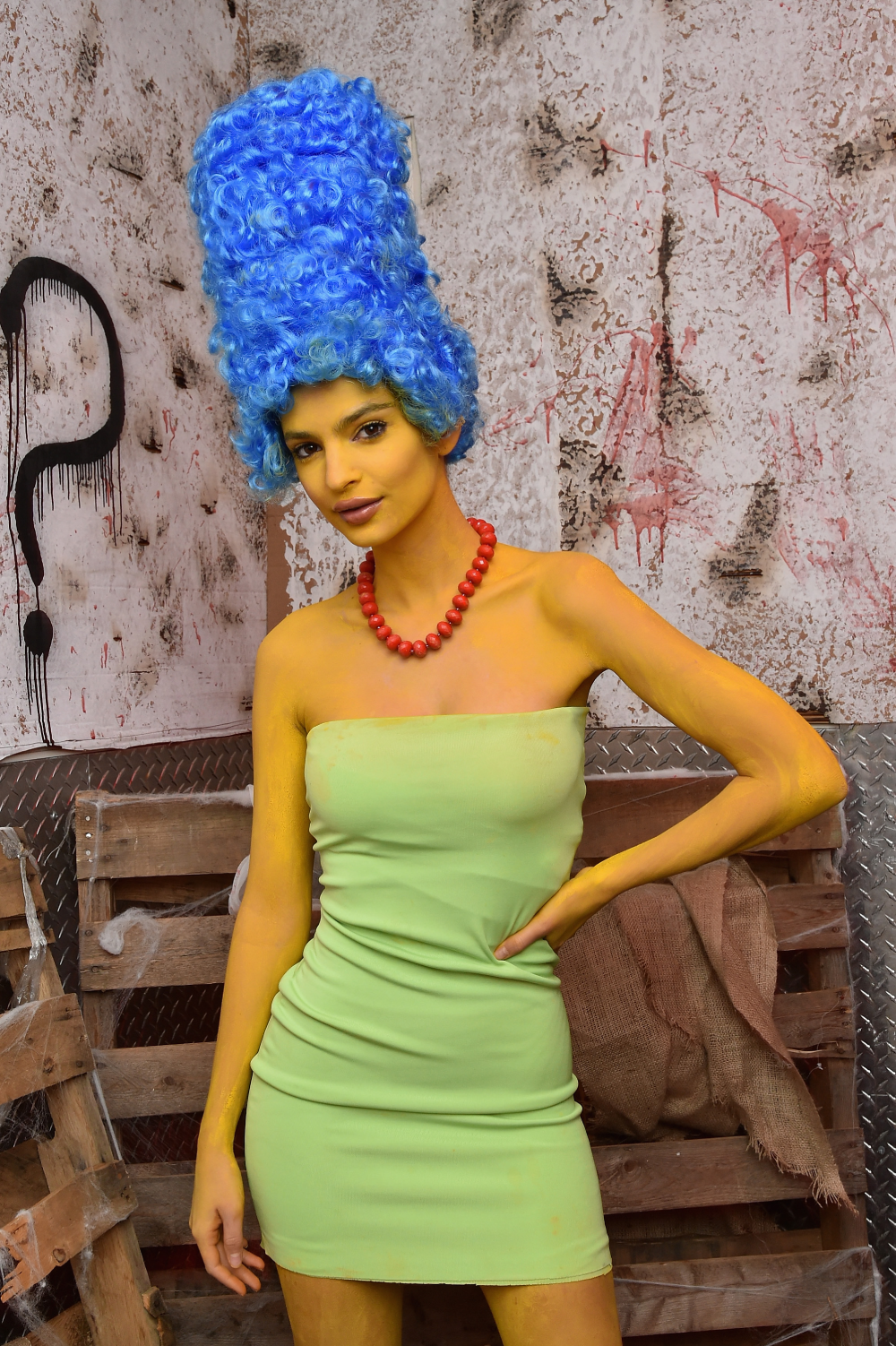 A woman in yellow body paint, a tall blue wig, and a red beaded necklace wears a green strapless dress as a Marge Simpson costume.