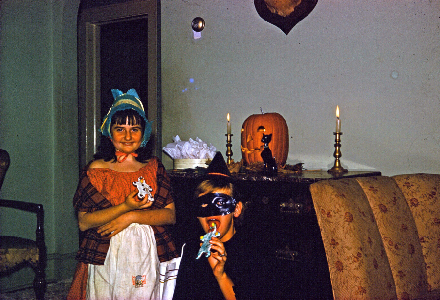 Two children in costumes celebrate Halloween indoors. One is dressed as a maid, holding a doll, and the other wears a mask, eating candy. A jack-o’-lantern and candles sit on a dark wooden dresser behind them.