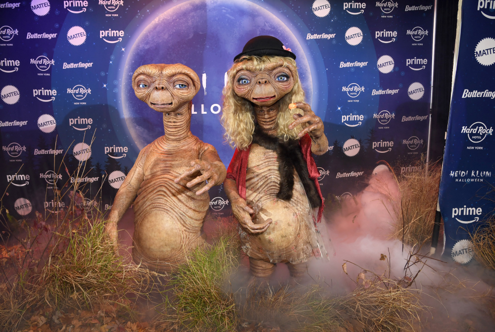 Two E.T. figures stand in tall grass amid ground fog in front of a branded backdrop.