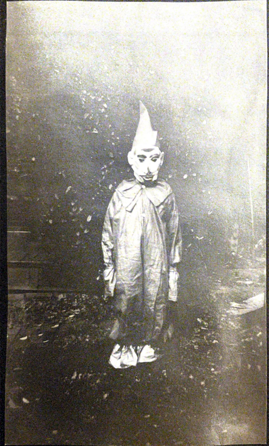 A person in an oversized clown costume and pointed hat stands outside. The mask has exaggerated features, and the photo is black and white with a faded, vintage appearance.